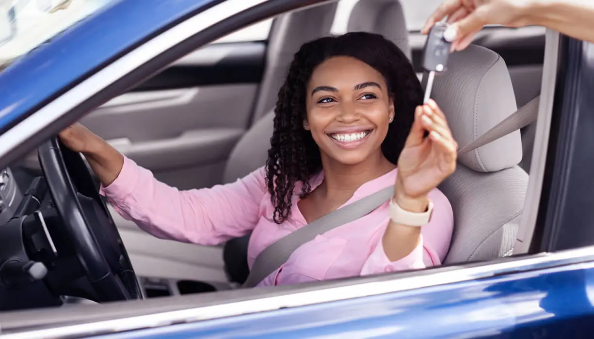 A smiling woman sits in the driver's seat of a car, hands on the steering wheel, ready to drive.