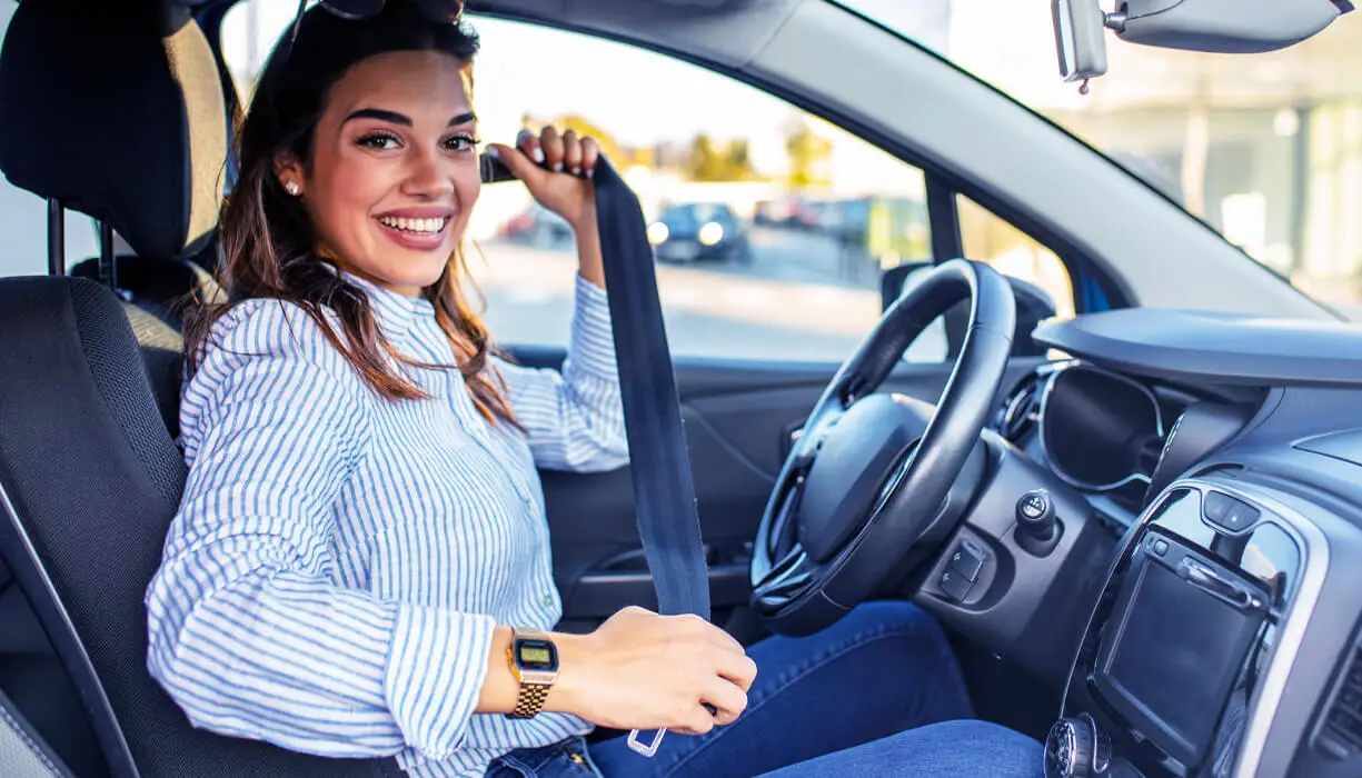 A young woman sitting in her car putting on her seat belt.