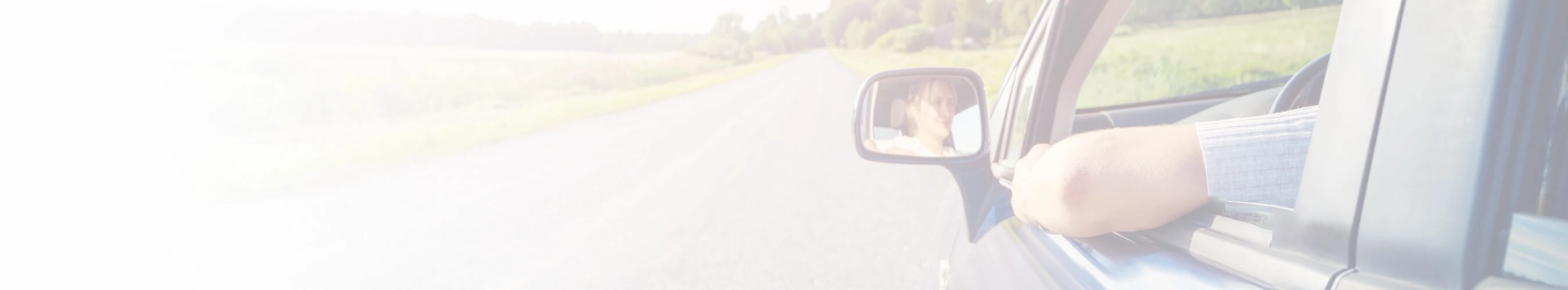 Person driving with their arm resting on the car window, looking at the road ahead with a view of the side mirror reflecting their face