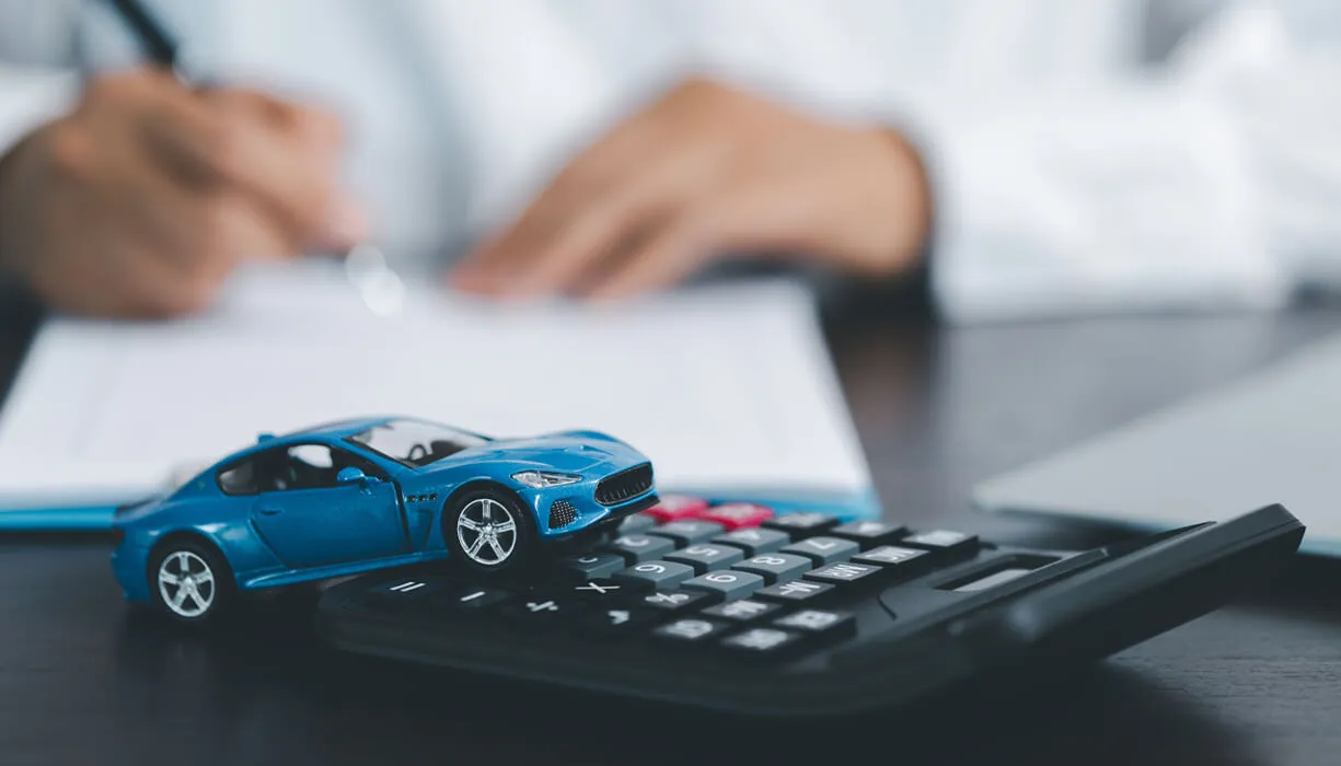 Close-up of a small blue toy car on a calculator, with a person in the background writing